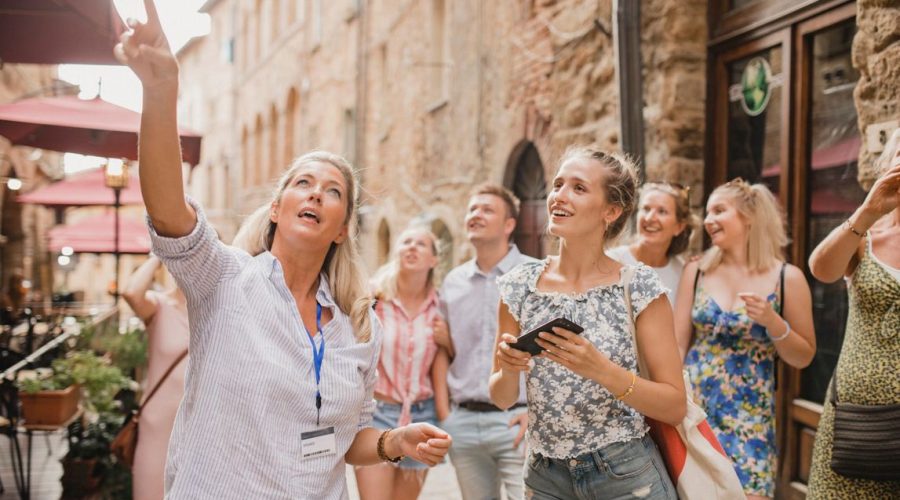 A tour guide gestures while speaking to a group of people on a narrow street lined with historic buildings and outdoor cafes, sharing insights that have earned rave tour reviews.