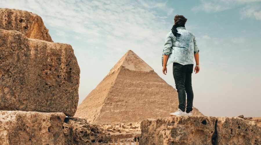 A person stands on a rocky ledge, facing the Great Pyramid of Giza under a partly cloudy sky, perhaps contemplating tour guide tipping while reading tour reviews.